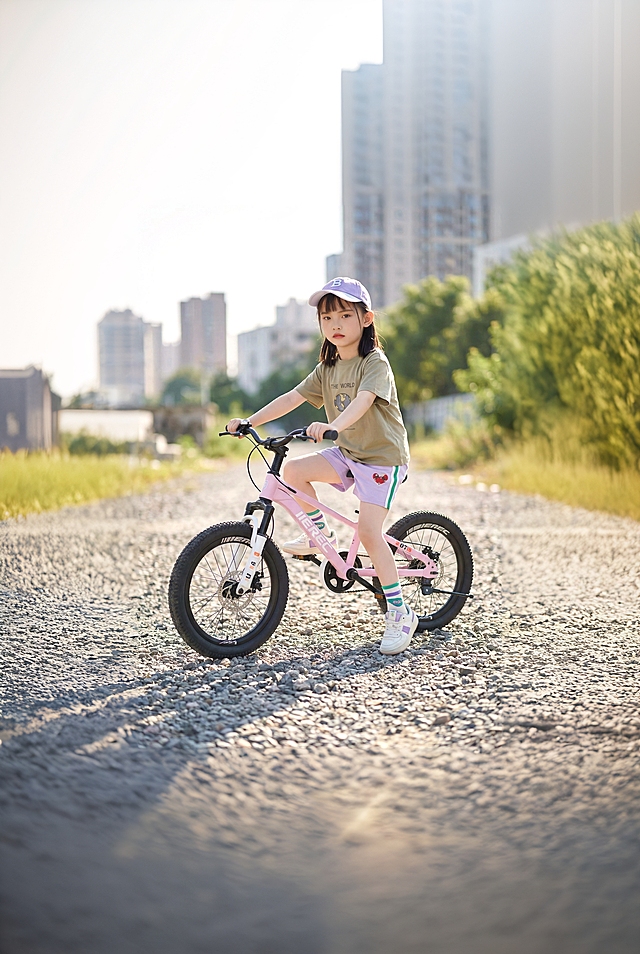 一个戴着浅紫色帽子的小女孩，穿着卡其色T恤和主色调为紫色侧面绿白相间的竖条纹，正骑在一辆粉色儿童山地车上，停在铺满碎石的户外小路上，背景是模糊的城市建筑和绿色植被，阳光明媚，整体风格自然清新，采用35mm焦距写实摄影，侧逆光拍摄，车身金属质感与碎石路面纹理清晰，色调以米白、军绿、浅灰为主，画面生动活泼充满童趣。