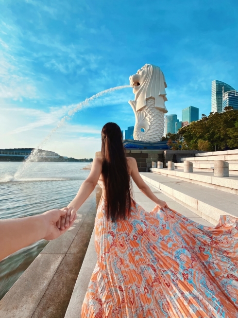 参考这张图片的风格和构图，重新生成新加坡鱼尾狮公园的牵手旅行照片，保留背成长发女生、橙色花纹长裙、牵手的手和鱼尾狮喷水的画面，保持明亮清晰的户外摄影风格，蓝天水域的场景。预览效果