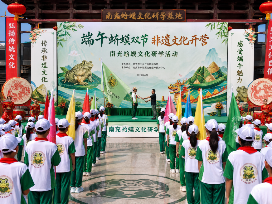 南充蛴蟆文化研学活动，非遗传承小队授旗仪式场景，背景为研学基地广场，人们身着研学服装，手持旗帜，主题为端午蛴蟆双节非遗文化开营，场景充满传统文化氛围预览效果