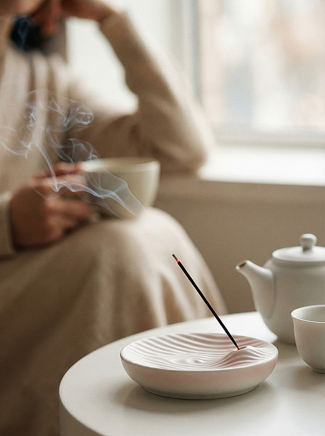 冬日午后的室内雅致角落，场景聚焦于靠窗的浅米色圆桌区域 —— 柔和的散射自然光从窗边漫入，带着淡暖的光晕包裹空间。画面核心是中下偏右位置的浅白渐变淡粉陶瓷香座：香座表面带有细腻起伏的波浪肌理，温润釉面泛着极淡的光泽，一支细长的黑色线香斜插其上，燃着的端头隐有微弱红火星，淡青灰色的轻烟正轻盈舒缓地蜿蜒上升，烟缕带着自然的柔卷纹理，既不浓郁也不飘散，质感轻盈而舒展。香座的右后方，置一套哑光白陶瓷茶具：茶壶壶嘴弧度柔和，小巧圆口的茶杯紧挨其侧，茶具表面晕着浅淡的环境反光，呈现出温润的哑光质感。
构图采用浅景深特写，焦点精准锚定线香与香座（细节锐利清晰），茶具处于中景层，仅带轻微虚化；背景则是极度模糊的远景：只能隐约瞥见淡米色宽松衣物的轮廓，裹着松弛倚靠的身形，手臂自然弯曲、手持的浅色杯子彻底融入虚化里，人物周遭的织物（沙发或靠垫）也只剩柔糯的色块过渡，全程以极弱的存在感点缀氛围，不抢任何视觉重心。
整体色彩是低饱和的暖柔色系，以暖白、浅米、淡棕为基底，佐以香座的极浅粉调，无生硬明暗对比，阴影是柔和的浅灰晕染。风格是东方极简美学，质感细腻却不张扬 —— 陶瓷的温润、烟雾的轻透、织物的柔糯皆以细微触感体现，氛围是冬日独处的慵懒松弛，安静舒缓里裹着淡淡的暖意，简洁又透着高级的雅致感。
