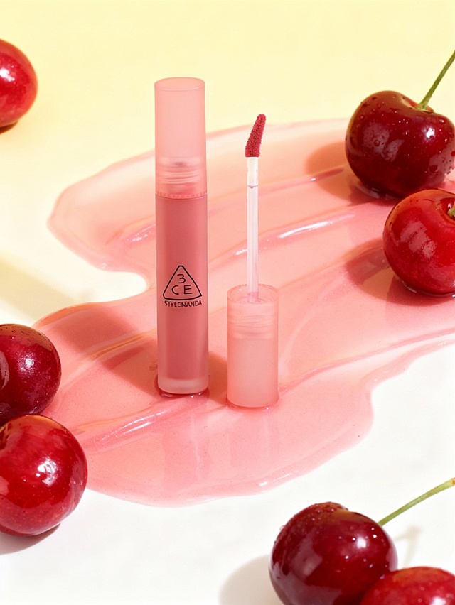 甜美水润美妆商业摄影风格，清新甜美的调性，画面整体采用粉红与浅黄渐变的暖柔配色，明亮柔和的柔光色调；采用俯视平拍视角，主体居中式构图；场景为浅黄至浅白的渐变纯色背景，前景是流动质感的粉色液态载体，搭配新鲜红樱桃元素；主体位于画面正中央区域，呈垂直竖立状态（旁侧放置打开的配件），占画面约 30% 比例，放置在粉色液态载体的表面；主体与环境的关系为：主体立于粉色液态载体之上，周围红樱桃环绕分布，液态载体的流动质感与樱桃的新鲜光泽形成呼应，浅黄渐变背景突出主体，明亮柔光布光强化水润甜美的整体质感，营造出清新甜美的美妆产品氛围。