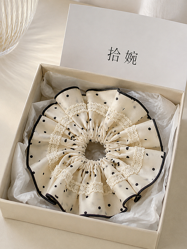 俯拍平铺静物摄影，韩系蕾丝波点大肠发圈（米白底黑色波点+白色蕾丝花边，面料刺绣纹理清晰细腻）放置在米黄色可降解纸浆盒内，盒内衬白色薄棉纸；盒子后方摆放白色纸质卡片，卡片印有黑色文字："拾婉"；干净浅米色桌面，柔和窗边自然光，韩式简约饰品静物，低饱和莫兰迪色调，高清电商实物实拍，浅景深，画面简洁高级