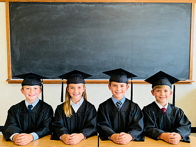 四位美国白人儿童毕业场景，在教室内正面朝向镜头拍照，后方是一整块没有任何字迹的大黑板，画面质感清晰锐利，细节丰富，色调自然明亮，呈现真实的毕业拍照氛围