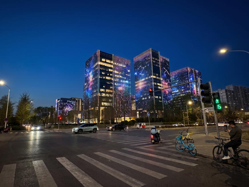 将这张白天的城市照片修改为专业的夜景照明方案效果图，保持建筑整体结构和布局不扭曲变形；着重突出画面中建筑玻璃幕墙的竖向结构肋条，在每个竖向结构肋条内安装彩色可变幻的线条灯具，所有竖向肋条上的线条灯组合预览效果