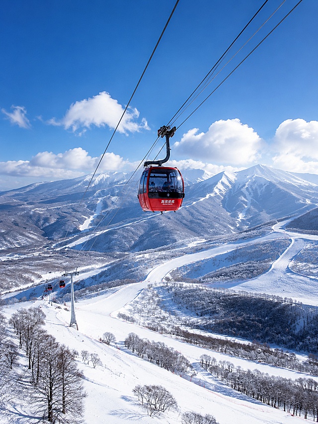 亚布力二锅盔山缆车索道冬季雪景，平视偏仰视视角，红色的缆车轿厢悬挂在钢索上，缓缓从雪山上驶过，下方是纵横交错的滑雪道和白茫茫的雪坡，远处是连绵的二锅盔雪山群峰，天空湛蓝，云朵漂浮，缆车玻璃反射着阳光，充满冬日度假的悠闲氛围，高清写实摄影，4K画质