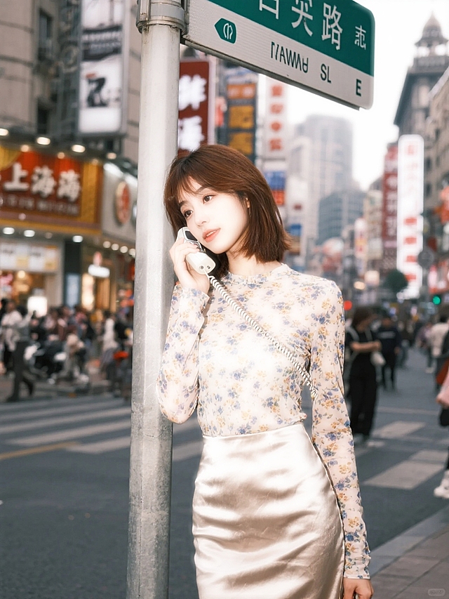 2000年的上海商场，繁华热闹，女生（碎花修身长袖搭配紧身绸缎半身裙，斜刘海发型）靠在路牌上，手拿小灵通电话。