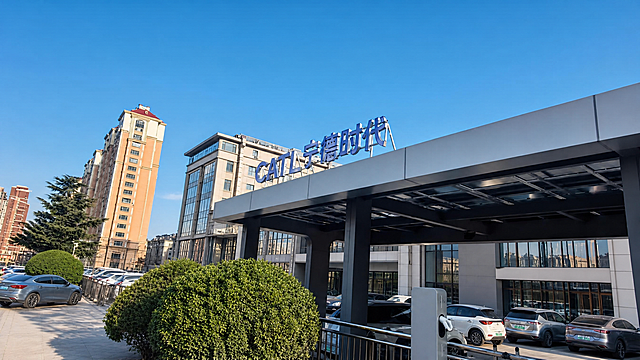 基于这张宁德时代场站实拍图进行画面整理优化：1. 去除画面左侧那道斜着的白色长杆和左下角的路灯杆，让画面更干净；2. 保留场站主体和CATL宁德时代招牌的真实样貌，不改变结构；3. 背景的楼房和周边商铺建筑做质感提升，外墙更整洁干净，窗户更通透，整体建筑看起来更高级精致；4. 天空调整为更纯净的湛蓝色，光线更明亮通透；5. 前景绿植、车辆、地面适当优化整洁度，整体画面更清爽有质感，色调统一高级，保留真实场景感，不添加任何文字，不要失真。