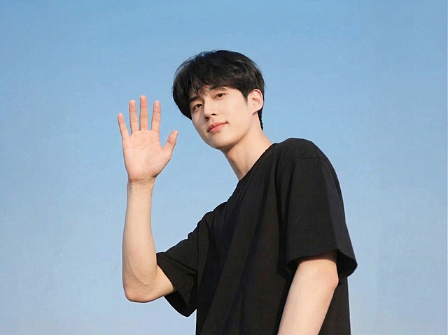 将图中男生原本放在额头旁的手势修改为挥手打招呼的姿势，保持人物其他特征、穿着和蓝天背景不变，自然真实