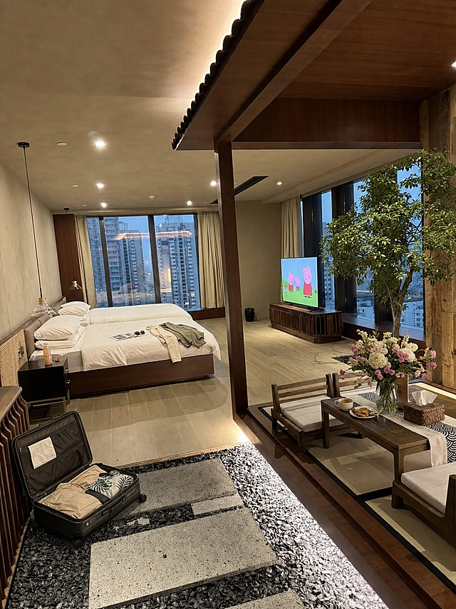 保持房间原有布局、家具、灯光和整体风格完全不变，进行以下修改：1. 去掉床上的两个小孩，恢复完整整洁的白色床品；2. 床上添加杂志、一副墨镜、一件搭放的夏季薄外套等生活物品；3. 地面角落放置一个打开的行李箱，箱内有夏季衣物；4. 茶桌上添加花瓶，花瓶里插着好看的鲜花，再添加咖啡杯、小食盘等生活物品；5. 右侧枯树枝上添加茂密的绿色树叶，变成生机盎然的绿树；6. 电视画面改为播放小猪佩奇动画片；所有物品透视和光影与房间环境完全一致，真实自然，摄影级写实