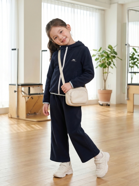 将image1中的藏青色连帽拉链外套和阔腿裤套装穿在image2的小女孩身上，背景为韩系休闲运动风格的高级专业摄影基地，搭配符合衣服风格的配饰，小女孩歪头甜笑表情，四分之三侧身展示，比例3:4预览效果