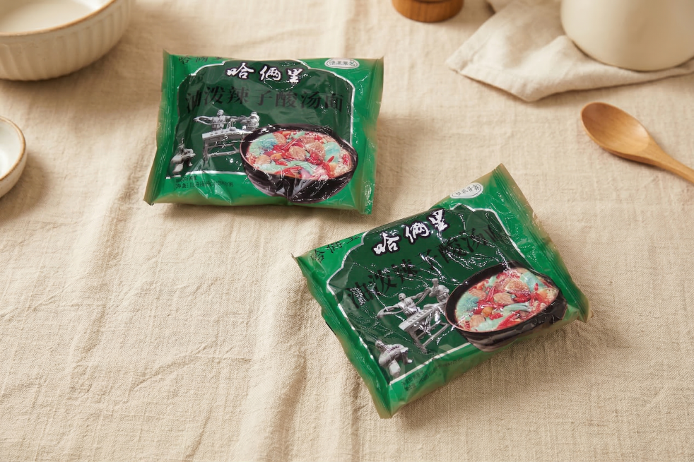 将两包哈俩里油泼辣子酸汤面泡面自然融入到铺有棉麻桌布的桌子上。1. 保持产品包装上的所有Logo和文字原样不变，保持主体物理结构不变，强制保留两个泡面包装袋完整组件。2. 光影匹配：采用全屋漫射柔光，预览效果