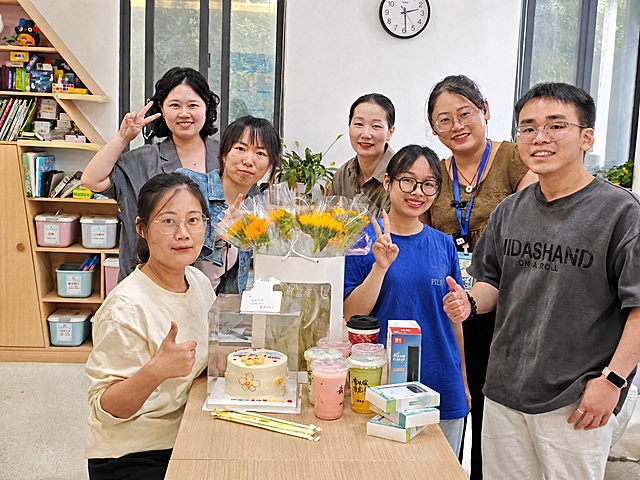 将第二张图中穿蓝色T恤戴黑框眼镜比耶的女生，自然地融入第一张集体合影中，站在队伍里合适的位置。要求：保留第一张图中所有人物、蛋糕、向日葵花束、饮品、礼物等全部元素和场景不变；新加入的女生外貌、服装、发型与第二张图完全一致；人物比例、透视角度、光影方向与第一张图完全匹配；边缘过渡自然，无拼接痕迹；整体画面协调统一，高清画质。