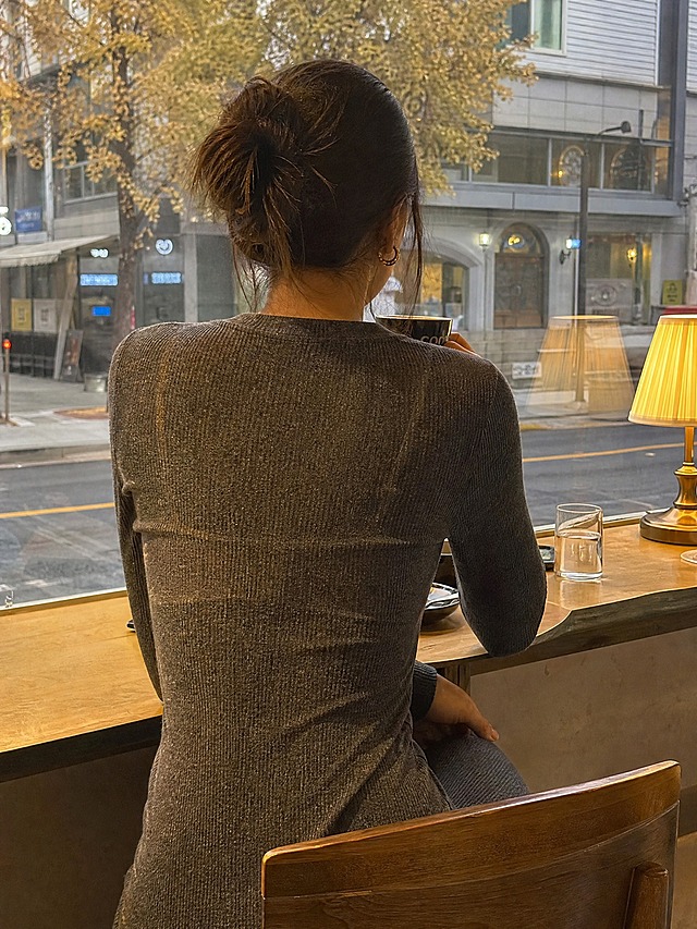 咖啡馆室内场景，从背后视角拍摄的女性坐在窗边喝咖啡，穿着深灰色厚实长袖针织连衣裙，裙装面料厚实不透明但剪裁修身，背部自然呈现内衣轮廓痕迹，肩带位置和罩杯形状透过针织面料隐约可见，秋季穿搭真实自然，不夸张不刻意，咖啡馆暖黄灯光氛围，画面写实高清，3:4比例