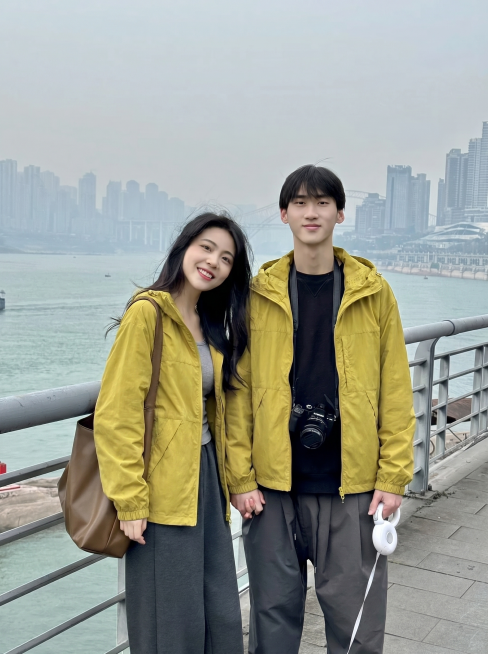 将图2的姜黄色连帽外套分别穿到图1江边护栏这对情侣身上，男女都换上这件姜黄色外套，替换掉原来的上衣，过渡自然平整，保持原来的江景城市背景和牵手姿势，图片比例3比4预览效果