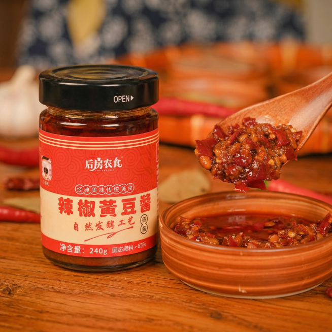 将这瓶辣椒黄豆酱生成美食商业摄影场景图，产品本体的形状、结构、图案、文字内容一字不改，文字清晰可辨，不模糊、不替换、不艺术化变形。酱料保留参考图本身的颜色、颗粒大小、酱豆形态、稠度、光泽，绝对不允许修预览效果