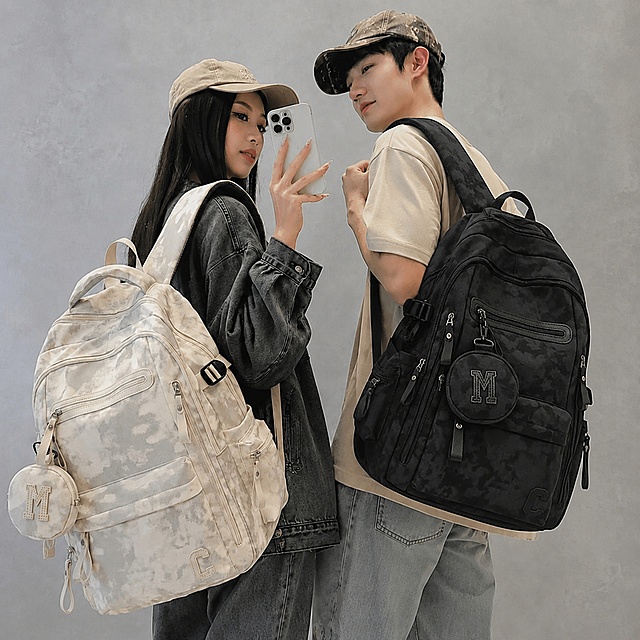 保留图中一对年轻情侣模特的姿势、服装、人物和背景不变，只将男生后背的背包替换为第二张图的黑色扎染双肩包，严格保留新背包的所有可见组件、轮廓、结构、比例、黑色扎染材质、颜色和M字样零钱包，保持光影和整体色调和原图一致，自然融合，不改变其他内容。