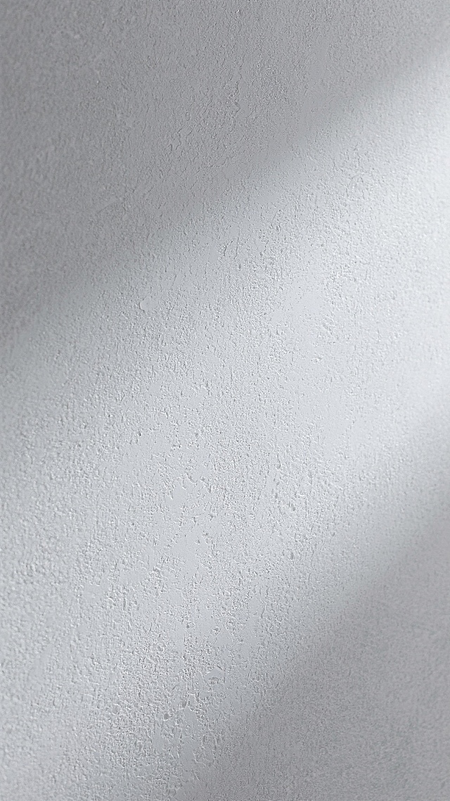 一张具有高度质感的灰色墙面背景图，墙面呈现出细腻的磨砂纹理，整体色调为浅灰色，带有轻微的明暗渐变效果，营造出简约而高级的视觉氛围。墙面材质看起来像是经过特殊处理的水泥或石膏，表面有自然的颗粒感和细微的凹凸不平，光线照射下形成柔和的阴影变化，没有任何文字、图案或装饰元素，纯粹展现材质本身的肌理美感。