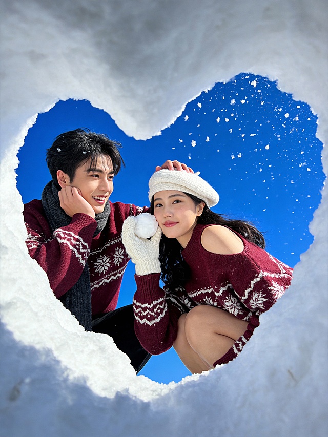极端的仰拍视角拍摄，以蓬松洁白的雪层挖空成的小心形框架为前景，从挖空的爱心学层从下往上的角度拍摄到外面的情侣，只看得到他们上半身，背景是高饱和度的纯净蓝天，同时添加动态飘落的细碎雪花，情侣靠近对方、头靠着头：左边男生穿酒红色底 + 白色北欧风雪花提花圆领毛衣，宽松版型，纹理清晰，搭配深灰色针织围巾，蹲着，一只手撑着下巴看女生，一只手抚摸女生的头，自然地开心笑着；右边女生同款酒红色底 + 白色北欧风雪花提花毛衣（做单侧露肩设计），宽松版型；白色针织贝雷帽。她侧身蹲着，回头看镜头，一只手带白色毛线手套，拿着雪球贴着脸，自然的微笑，两人身体贴在一起。画面采用极具冲击力的超广角鱼眼镜头“自拍”视角，整体光影为自然日光，色彩鲜艳，8K超高清画质。高饱和色彩，网红打卡感，营造冬日户外鲜活氛围感。