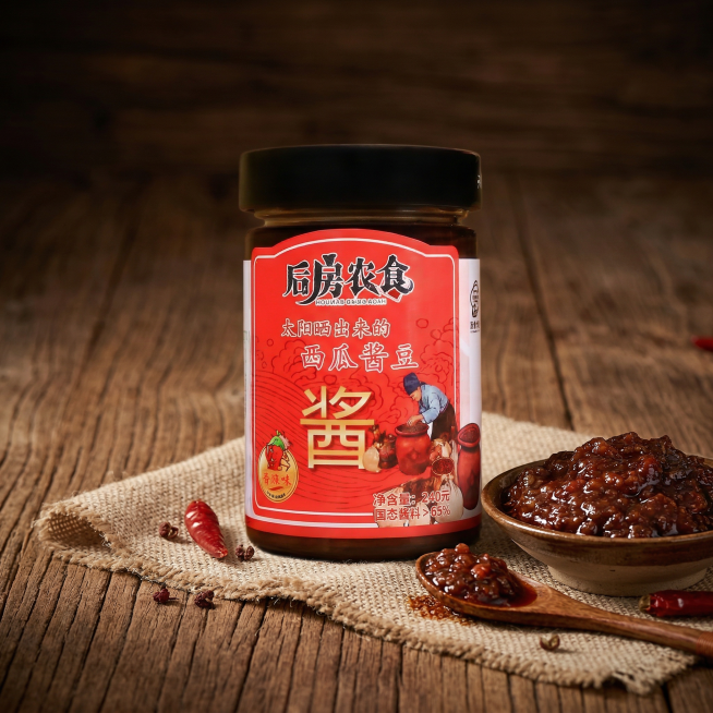 以图1的后房农食西瓜酱豆为主体,瓶身标签所有文字和细节完全保留不做修改。背景为深棕色复古实木桌面,粗粝木纹清晰,搭配麻布衬底,木勺、小瓷碟、辣椒作为点缀,小瓷碟中的酱体使用图2里深褐色油亮、颗粒饱满的预览效果
