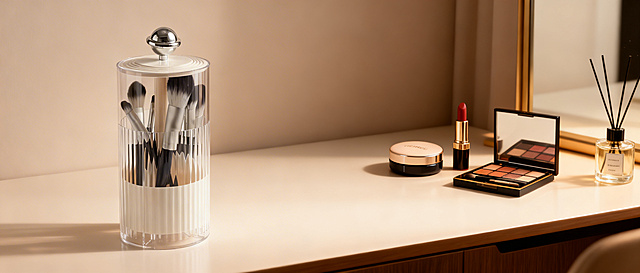 透明带盖化妆笔刷筒放在画面最左侧，背景为简约高级的梳妆台场景，右侧有气垫、口红、眼影盘和小香薰，柔和暖光，精致美妆氛围，突出笔刷筒防尘收纳的实用性