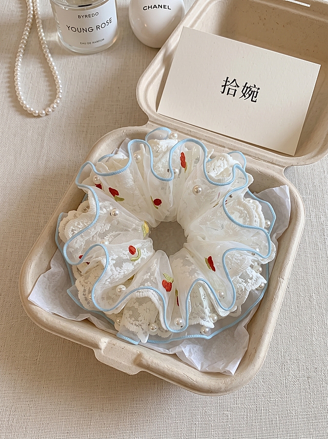 俯拍平铺静物摄影，白色刺绣花朵网纱蕾丝双层大肠发圈，带有红色樱桃刺绣、珍珠装饰、浅蓝色滚边，放置在米黄色可降解纸浆盒内，盒内衬白色薄棉纸；盒子后方摆放白色纸质卡片，卡片印有黑色文字"拾婉"；干净浅米色桌面，柔和窗边自然光，韩式简约饰品静物，低饱和莫兰迪色调，高清电商实物实拍，浅景深，面料刺绣纹理清晰细腻，画面简洁高级，3:4竖版构图