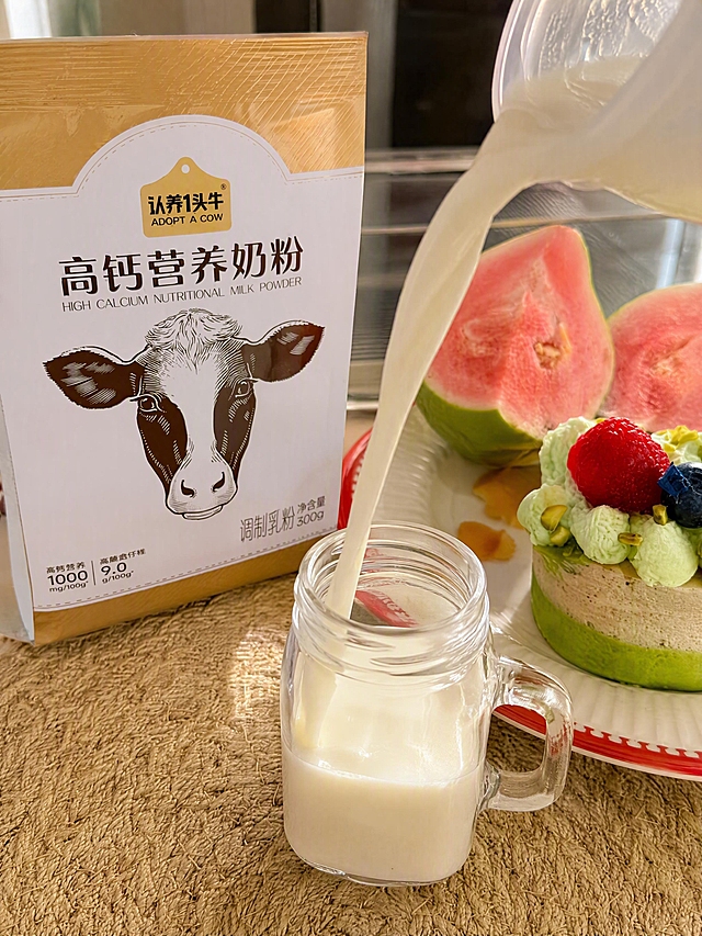 奶液冲调画面，乳白色泛黄奶液正注入透明玻璃小奶杯中，形成优美的流动弧线，旁边摆放认养一头牛高钙营养奶粉袋装产品，编织桌面，芭乐和小蛋糕点缀，真实手机拍摄质感，暖光自然光，生活感，3:4竖版构图