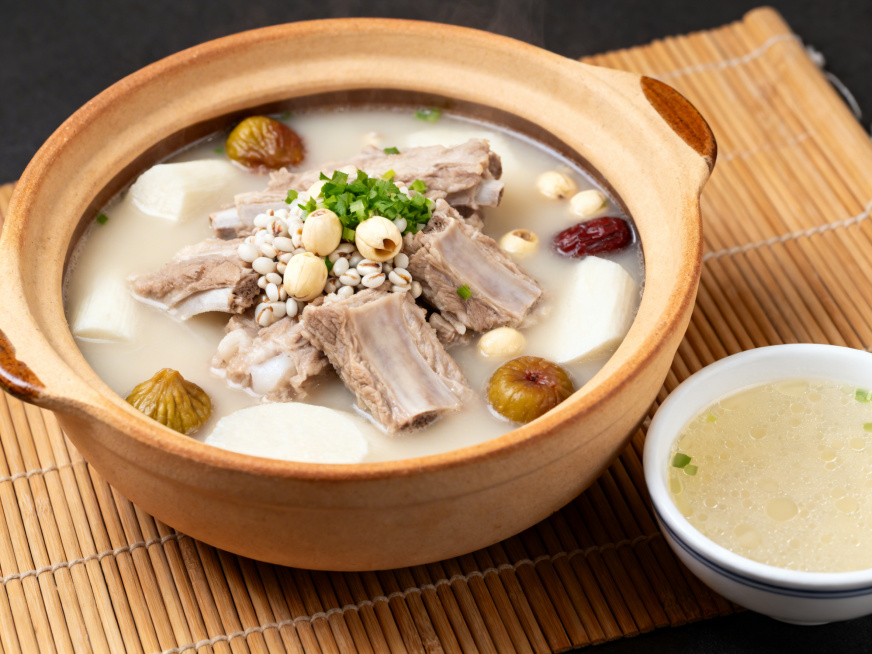 广式养生猪排汤，肋排炖至酥嫩脱骨，汤色清润呈浅奶白色，搭配莲子、薏米、山药、无花果等清补食材，撒上少许切碎的香芹碎提鲜，盛放在哑光陶制汤煲中，旁边摆一小碗盛出的汤，汤面飘着薄薄一层油花，摆放在竹制餐垫预览效果