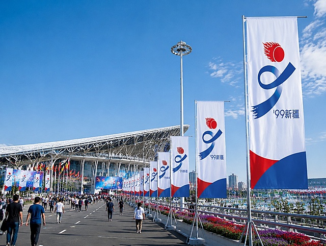 运动会道旗样机效果图，道旗上印有参考图中的运动会LOGO（红色火焰太阳+蓝色数字9造型+新疆字样），红蓝配色，多面道旗排列在宏大的运动会会场道路两旁，广角镜头，写实摄影风格，高清细节，专业样机展示，会场画面宏大壮观，有行人观众人物元素，真实光影质感