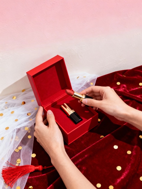 礼盒开箱图
 
核心提示词：俯拍视角，商业产品摄影，一双手打开红色方形礼盒，取出一支口红，盒内还有另一支口红，背景为浅粉色渐变墙面，搭配红色丝绒布、白色薄纱、金色亮片和红色流苏，整体氛围精致有仪式感，预览效果