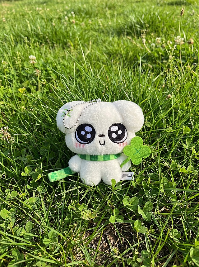 保留图中的白色玩偶挂件主体，玩偶可调整位置，将背景替换为真实的绿色自然草地，玩偶和背景融合自然，带有真实阴影