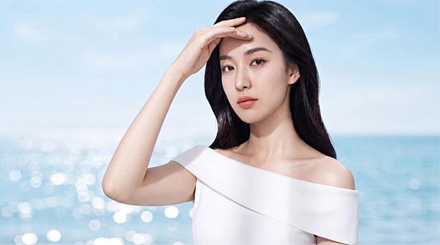 The beautiful Asian woman slowly raises her hand to gracefully shield her eyes from the glaring sun. She squints slightly but elegantly. The bright sunlight creates a beautiful glow on her skin. In the beautifully blurred background, the light-blue ocean sparkles and subtle waves move. Soft sky-blue color tone. High-quality commercial beauty shot.