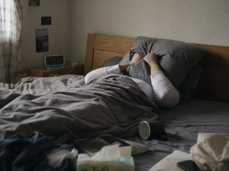 4:3构图，一个人躺在床上，盖着杯子，用枕头烦躁地捂住脸，床上物品有些杂乱，床头位置放着一个闹钟，生活化场景，自然光线，氛围沉闷预览效果