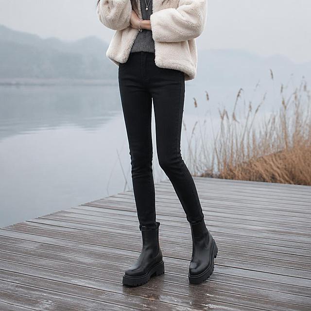 以参考图中的小脚牛仔裤为唯一服装主体，严格还原裤子细节：纯黑色、哑光牛仔面料（无反光、无油亮高光）、小脚裤型（裤腿自上而下自然收窄、紧贴小腿并在脚踝处收束）、面料质感、腰头、口袋与压线、缝线及垂坠感均保持原样。为这条裤子搭配一位全新模特，画面只展示腰部以下部分：从腰腹位置开始裁切，完整展示双腿至脚部，不出现头部、脸部与上半身。上衣更换为全新冬季搭配：奶白色短毛绒短外套（下摆自然盖住腰头），内搭深灰色高领针织衫；鞋子更换为全新冬季单品：黑色厚底切尔西短靴；另以银色细项链与银色细手镯作点缀，全部为全新搭配，不使用参考图中的原有上衣、鞋子与首饰。场景为韩系冬日湖边木栈道：干净木板栈道、两侧枯萎蒹葭与薄雾、冷色调平静水面与朦胧山影、清透柔和的漫射光，安静高级的韩系氛围；背景不得出现任何文字、招牌、图标、水印或品牌标识，也不得出现店铺、路牌或任何可辨识的文字图案。高档韩系商业时尚摄影，低饱和冷调，真实面料纹理与自然光影，8K 细节。
