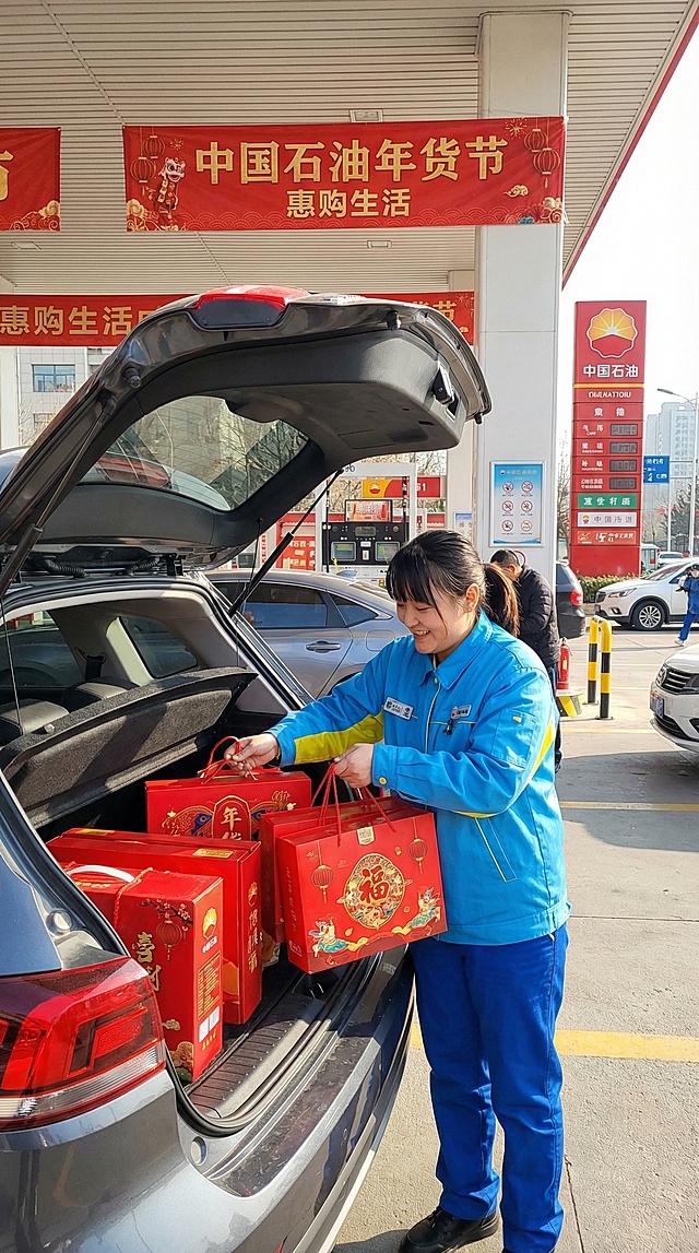 中国石油加油站内，一位身穿蓝色制服（参考参考图中的服饰））的加油员微笑的给汽车后备箱，搬放年货