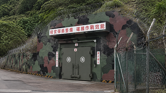 台军弹药库外景，钢筋混凝土结构，厚重防爆门，迷彩伪装，周围有铁丝网和警戒设施，山地隐蔽环境，写实风格，高清细节