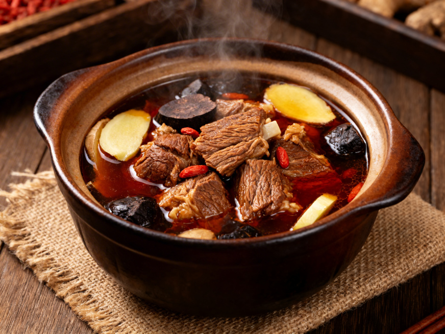 家常食补菜何首乌羊肉，大块羊肉炖至酥软入味，色泽红亮，搭配切片何首乌、枸杞、姜片等食材，汤汁浓郁醇厚，表面浮着淡淡油光，盛放在深褐色陶土炖锅中，锅边冒着微微热气，摆放在粗麻餐垫上，暖调光线照射下，羊肉预览效果