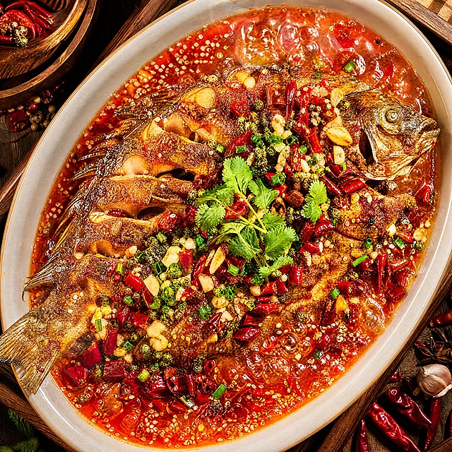 一张极具食欲的烤鱼美食摄影照片，完整的烤全鱼放置在椭圆形深瓷盘中，鱼身表面金黄焦脆带有漂亮的烤纹焦斑，淋满红亮的红油汤汁，上面铺满鲜红的干辣椒段、青花椒、蒜末、葱花和香菜，鱼肉微微裂开露出白嫩的内里，汤汁咕嘟冒泡冒着热气，盘边点缀着白芝麻和红油光泽，俯拍视角，暖黄色柔和灯光，高对比度，色彩鲜艳饱和，细节极其丰富，专业美食摄影风格，令人垂涎欲滴