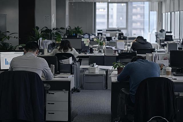 现代开放式办公室场景，多名员工穿着商务休闲装（衬衫、针织衫、休闲裤等），以背影、侧影、低头角度呈现，无需露出正脸，整体氛围士气低落压抑，有人趴在桌上，有人垂头靠在椅背上，有人无力地敲着键盘，桌上散落文件和咖啡杯，冷色调柔和光线，写实摄影风格，真实办公环境，高清细节