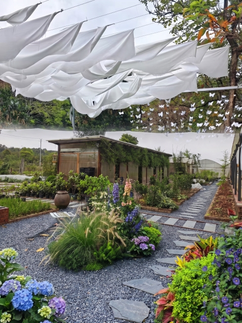 在图1（木质小屋+菜园式布局）基础上，融合图2的现代轻奢庭院风格进行升级：保留房屋主体结构、位置及周边砖砌花坛完全不变；将前景与中景的裸露土地与稀疏植物替换为图2风格的精致园景——包括修剪整齐的球形灌预览效果