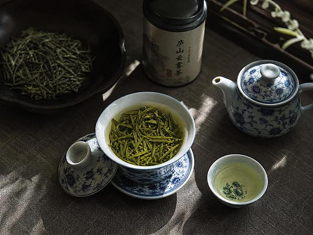 中式静物摄影，俯拍视角，复古粗麻灰色茶桌，青花瓷三才盖碗，盖碗内泡满嫩绿细长庐山云雾茶叶，清浅嫩绿茶汤，旁边搭配同款青花品茗杯，右侧摆放青花小茶壶，还有一小罐标明庐山云雾茶的小茶罐，背景深色木盘盛放干茶叶，窗边柔和侧逆光，光影斑驳，暗调氛围感，低对比度，素雅新中式，写实高清，胶片质感，景深虚化背景，画面静谧治愈，茶文化静物，8K，细节细腻