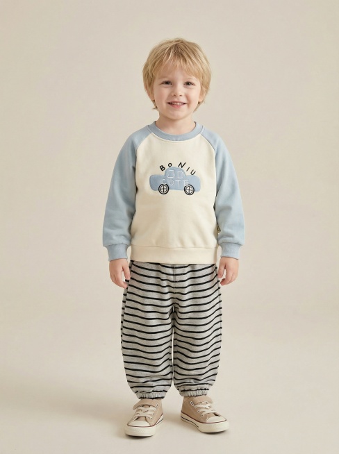 生成3岁欧洲男童童装目录封面图，男童为浅肤色、自然软发、面带温暖自然的微笑，摆出自信帅气的站姿；穿着上传的米白拼浅蓝插肩袖汽车刺绣卫衣、黑灰条纹束脚灯笼裤（调整为及踝长裤型），搭配米白色匡威帆布鞋；背预览效果