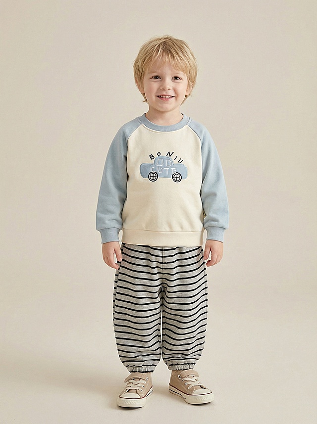 生成3岁欧洲男童童装目录封面图，男童为浅肤色、自然软发、面带温暖自然的微笑，摆出自信帅气的站姿；穿着上传的米白拼浅蓝插肩袖汽车刺绣卫衣、黑灰条纹束脚灯笼裤（调整为及踝长裤型），搭配米白色匡威帆布鞋；背景为浅米色极简风格，采用柔和自然光线，低饱和度中性配色，突出服装质感细节，整体氛围优雅干净温暖自然，无文字