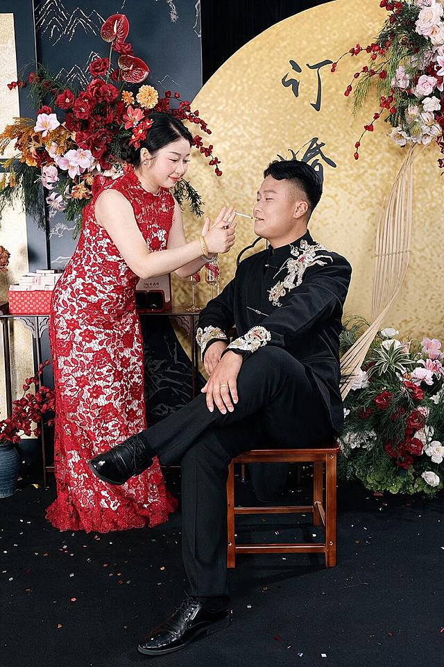 超高清8K分辨率，RAW原片画质，专业人像精修，全身构图新中式订婚写真，喜庆纪实氛围，东亚年轻情侣互动场景；女士站立俯身，身着红白蕾丝无袖长款改良旗袍，低盘发点缀红色花朵发饰，佩戴珍珠耳钉与金手镯，双手为男士点烟，神态温柔带笑；男士翘二郎腿坐在实木方凳上，身着胸口与袖口带有银白龙纹刺绣的黑色立领新中式礼服，脚穿黑色亮面皮鞋，神态放松自然。背景为新中式订婚布景：金色圆形背景板印有黑色书法汉字，左侧深色山水纹背景板搭配红掌、玫瑰、雏菊组成的高低错落花艺，旁置中式条案，摆放红色礼盒、喜酒与高脚杯，右侧搭配粉红花艺与竹编装饰，地面为黑色地毯，散落彩色亮片碎纸。采用中性灰分层磨皮、高低频分离修图，保留完整皮肤毛孔与真皮纹理，去除面部瑕疵、痘印、细纹；局部光影重塑，双曲线光影过渡，面部光影均匀化；液化遵循人体骨骼结构，颅顶自然优化、下颌线柔和收窄，五官微雕处理：眼型自然微调、鼻梁立体度强化、唇形精致修型；色温5600K自然白光，低饱和肤色校准，面颈肩肤色统一无色差；暗部提灰保留完整细节，高光压制无过曝爆边，镜头暗角轻微弱化；采用USM智能锐化，边缘锐化克制，整体画面通透干净