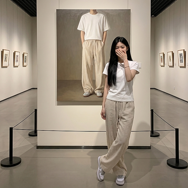 一位清冷气质的18岁东亚年轻女生，留着黑长直发型，身穿纯白色短袖上衣，下身搭配米白色亚麻休闲阔腿裤，脚踩浅灰色耐克板鞋。她身后是一幅等身大正方形的男性写实油画，画中人物穿着同款上衣和同款亚麻休闲裤，双手插兜，与女生形成呼应式穿搭。展厅两侧墙面挂有小幅艺术画作，地面设有黑色隔离立柱，女生右手捂嘴浅笑看向镜头，微微侧身但是不露脸，双腿轻交叉，整体氛围清冷高级，兼具艺术感与穿搭展示性，光线柔和且层次分明，突出人物与画作的互动感