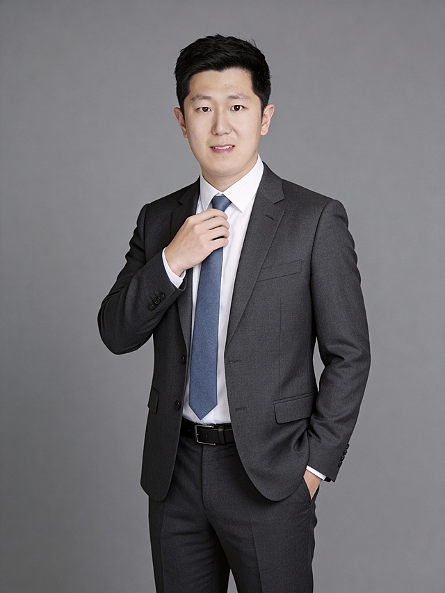 参考第一张图保留人物脸型､发型､年龄特征生成职场形象照,正脸照;人物居中,消除痘印,肤质优化处理提亮肤色,均匀肤色,干净的男士淡妆,气色自然,无明显浓妆痕迹;身穿暗灰色西装西裤,白色衬衣,灰蓝色领带,暗色皮带,西装剪裁合身,面料挺括,一手轻调整领带,另一手插于裤兜,站立,臀部以上半身像,表情动作自然,眼神坚定直视镜头,表情沉稳淡然;淡妆,柔和自然打光,画质清晰,头部完整,高清棚拍专业摄影人像写真,去除人物之外的所有元素,50MM标准焦段,F/4光圈(F/2.8-F/4区间),相机与人物距离2米,横向16:9比例(左右预留环境空间),人物占画面60%,入镜头部至膝盖上方20CM,平视视角,头肩比例协调,结构准确;光比1:1.5,4800K中性光,修饰脸型,肤质哑光细腻,面部雾面柔光,轻透裸肌感,真实毛孔纹理,32K画质,去除人物之外的所有元素,零噪点降噪,中深灰度纯色背景｡