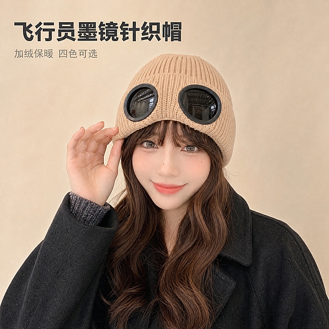 [主图-1] 飞行员墨镜针织帽正面上头效果展示，突出帽型与整体佩戴效果。
[统一基调] 1688源头工厂品质风，简约干净，浅米色肌理背景，主标题深灰粗黑体，辅助文字浅灰色，整体柔和时尚摄影光，高级松弛感。
[画面] 年轻亚洲女性模特，棕色长卷发，精致淡妆，面带自然微笑，胸部以上近景构图，完整展示帽顶与帽檐，不裁切头部。模特佩戴奶茶卡其色飞行员墨镜针织帽，帽子正面朝前，高清黑色圆形墨镜镜片清晰可见，针织纹理细腻，翻边设计工整。模特身穿黑色毛呢大衣，单手轻触帽檐，姿态自然松弛。背景为浅米色纯色墙面，简洁干净无杂物，突出帽子主体。帽子占画面主体视觉中心，大小适中，清晰展示帽型、颜色和墨镜镜片细节。
[光影质感] 柔和时尚摄影光，主光从左前方45度打来，面部光线均匀，帽子针织纹理有自然明暗过渡，墨镜镜片有轻微环境反光，质感真实，阴影柔和不生硬。
[配色] 整体暖米色调，帽子奶茶卡其色为主，黑色墨镜镜片和黑色大衣形成对比，背景浅米色，文字深灰色。
[文字版式] 左上角放置主标题「飞行员墨镜针织帽」，深灰色粗黑体大号字；下方小字「加绒保暖 四色可选」，浅灰色细体。文字不遮挡帽子主体和人脸。
[输出要求] 1:1正方形比例，2K分辨率，文字清晰可读，产品清晰不变形，无水印无平台Logo，无多余杂物。