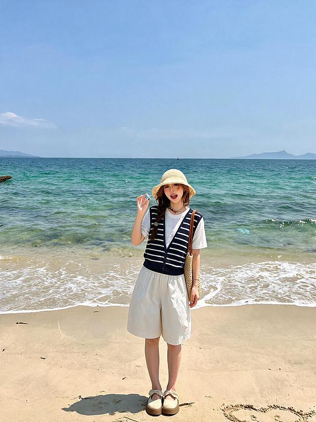 竖幅3:4，手机原相机直出纪实海边人像照片，年轻东亚元气少女，单侧麻花长辫，头戴草编宽檐遮阳草帽，活泼甜美的表情，一手抬起拿着小饰品，海边松弛站姿。身穿黑白条纹假两件针织短袖上衣，V领条纹针织，薄款透气冰丝针织面料，清晰细密针织网眼肌理，面料柔软带有细微颗粒肌理感，轻盈微垂，下摆罗纹收边。下身米白色茧型宽松五分中裤，哑光微光泽聚酯纤维面料，裤型宽松蓬量饱满呈茧型弧度，裤长至膝盖中间位置，4cm宽高腰松紧腰头，腰头两侧有细密竖向褶皱自然堆叠，裤身带有弧形分割缝线，两侧弧形插袋，裤腿宽大圆润自然垂落。脚上穿着米杏色蝴蝶结玛丽珍包头半拖，磨砂绒面材质，厚棕色生胶底。单肩挎镂空草编托特包，草编镂空肌理清晰，搭配棕色皮质长肩带。背景是干净的海边沙滩，清澈渐变青蓝色大海，远处朦胧山峦，晴朗淡蓝色天空，海浪轻轻冲刷沙滩，明亮通透的日间自然光，清新夏日海滨度假写真，原生真实皮肤肌理，衣物面料纹理清晰可见，高清写实，清爽治愈氛围感