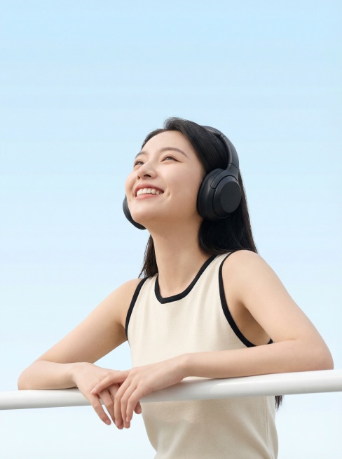 一位年轻的东亚女性，身穿米白拼黑边的无袖紧身背心，戴着黑色头戴式耳机，手肘搭在一根白色横杆上，身体微微前倾，仰头看向画面左上方，露出愉悦的笑容。背景是纯净的渐变浅蓝色天空，整体光线明亮柔和，画面构图简预览效果