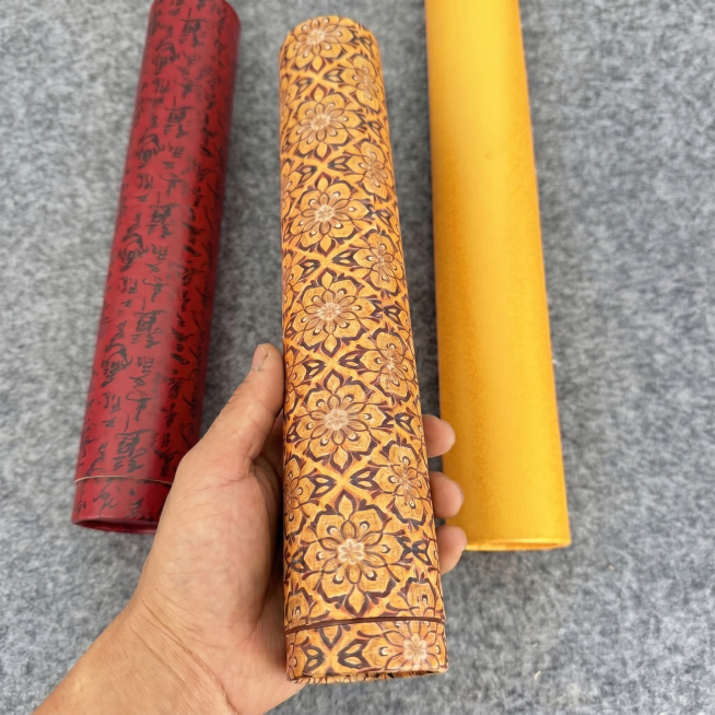 将这张图最左侧的橙色纯色画筒替换为图2中间的红底带黑色书法纹样的画筒，保留原产品红色底和黑色花纹，保持手的姿势、灰色背景、中间带花黄筒和右侧黄筒不变，光影自然预览效果