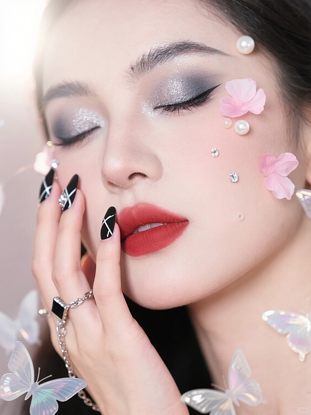 一张特写人像摄影，柔和的光线从左上方照射，营造出温馨梦幻的氛围。画面中女性闭着双眼，面部肌肤白皙细腻，脸颊和眼角点缀着精致的粉色花瓣、小巧的珍珠和闪亮的水钻装饰，形成自然散落的美感。她的妆容变换为冷调烟熏妆，眼窝处用深灰色和银色眼影晕染，眼线细长上扬，睫毛浓密卷翘，唇色改为哑光正红色，凸显冷艳气质。她的手轻轻托着脸颊，指甲变换为黑色磨砂底搭配银色几何线条和菱形水钻的美甲样式，指尖还点缀着小巧的金属链条装饰，整体造型融合了冷艳与精致的风格，背景中散落着透明的蝴蝶装饰，增添灵动飘逸感，画面整体色调在保持柔和光线的同时，通过妆容和美甲的冷色调形成对比，突出变换后的妆造主题。
